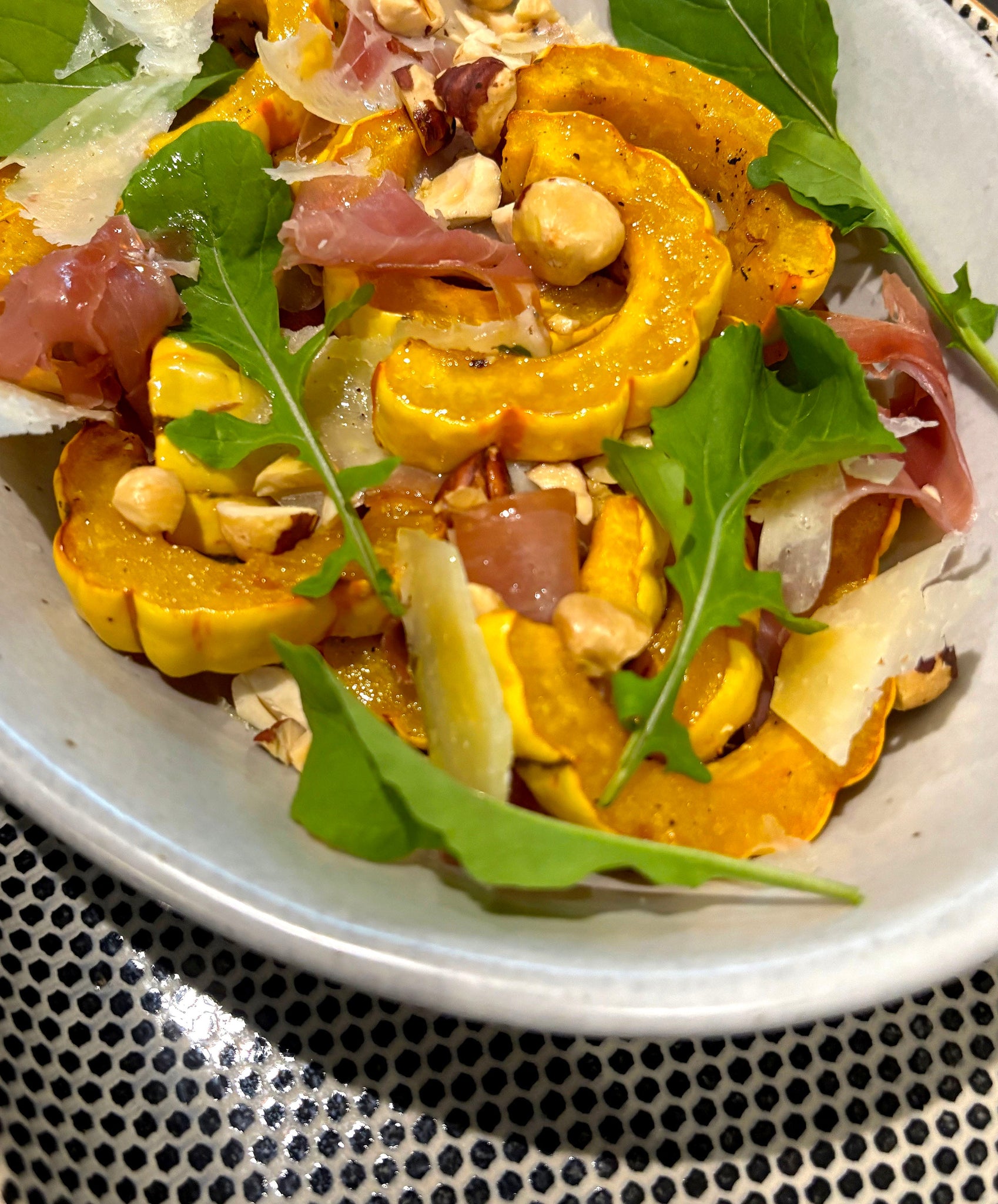 A close up of a  plate of sliced, roasted delicata squash, plated with shallows, prosciutto, hazelnuts, and arugula.